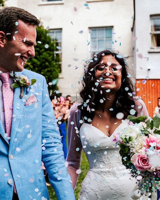 The bride and groom smile as they are showered with confetti outside at No.4 Clifton Village, a unique wedding venue in Bristol with a garden. The bride, holding a vibrant bouquet of pink roses and white flowers, is beaming with happiness, while the groom, in a light blue suit, shares in the joyful moment. The wedding venue garden, with its lush greenery, serves as a picturesque setting for this intimate wedding celebration, showcasing the venue’s charm and suitability for outdoor weddings.