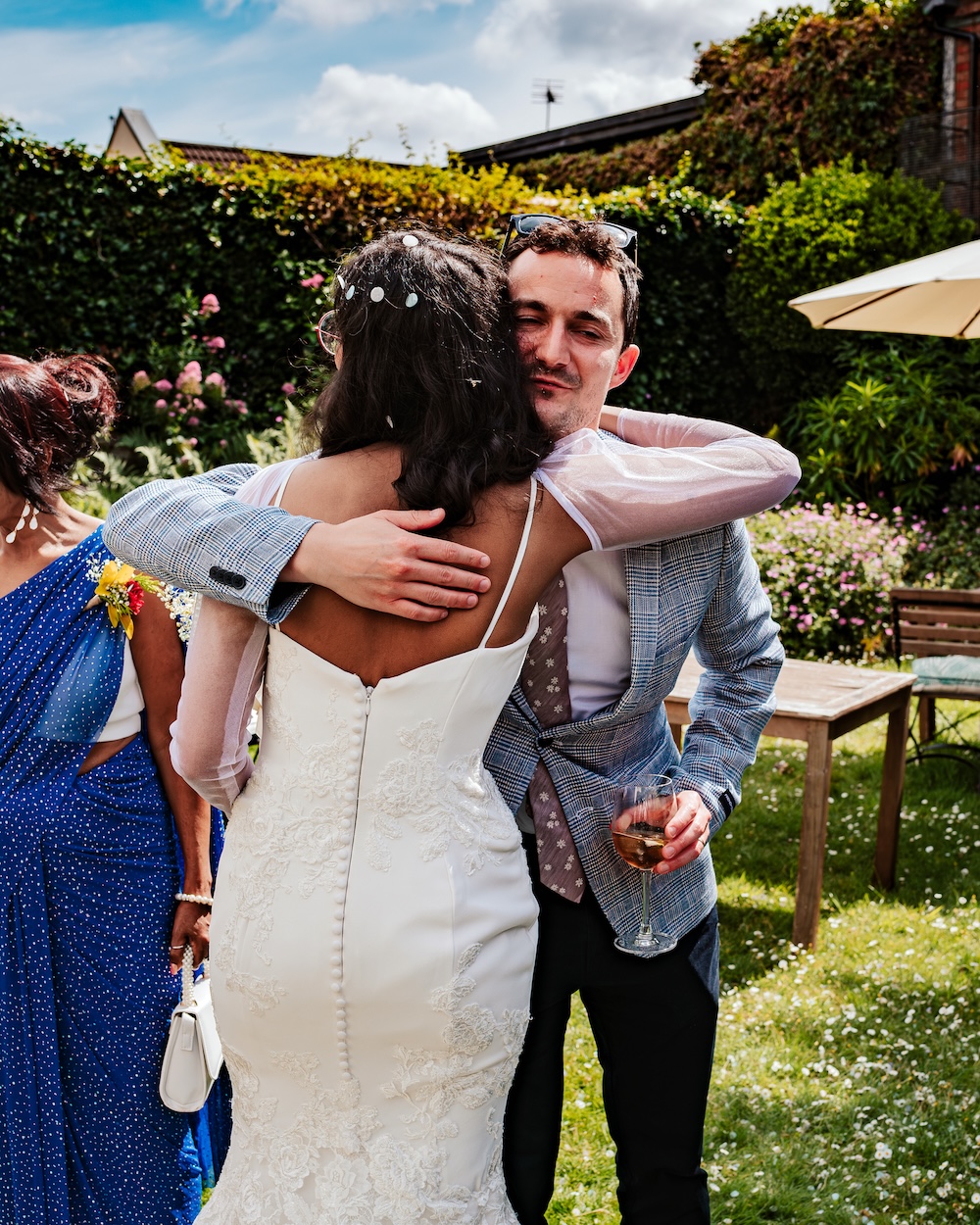 The bride embraces a guest warmly in the garden of No.4 Clifton Village, an intimate wedding venue in Bristol. The back of her elegant white lace dress is visible as she shares a moment of joy with the guest, who holds a glass of wine. The garden at No.4 Clifton Village, with its vibrant flowers and greenery, provides a relaxed and welcoming atmosphere, perfect for celebrating with close friends and family in this unique wedding venue.