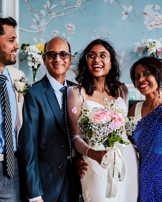 A happy family moment captured at No.4 Clifton Village, a boutique wedding venue in Bristol. The bride, holding a bouquet of pink and white flowers, stands with her family members, all smiling brightly. The elegant blue wallpaper with floral designs in the background adds to the intimate and sophisticated setting of this unique wedding venue. This image reflects the warm and personal nature of the venue, ideal for couples looking to create cherished memories with their loved ones.