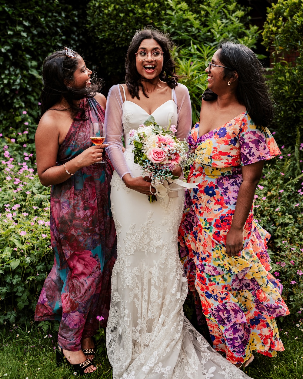 A candid moment captured at No.4 Clifton Village, a renowned Bristol wedding venue, shows the bride standing with two bridesmaids, all sharing a laugh. The bride, holding a bouquet of pink and white flowers, looks surprised and delighted, while her bridesmaids, dressed in vibrant floral gowns, join in the laughter. The lush greenery and pink flowers in the background emphasize the garden’s natural beauty, making this Clifton wedding venue a top choice for couples desiring an outdoor, intimate setting.