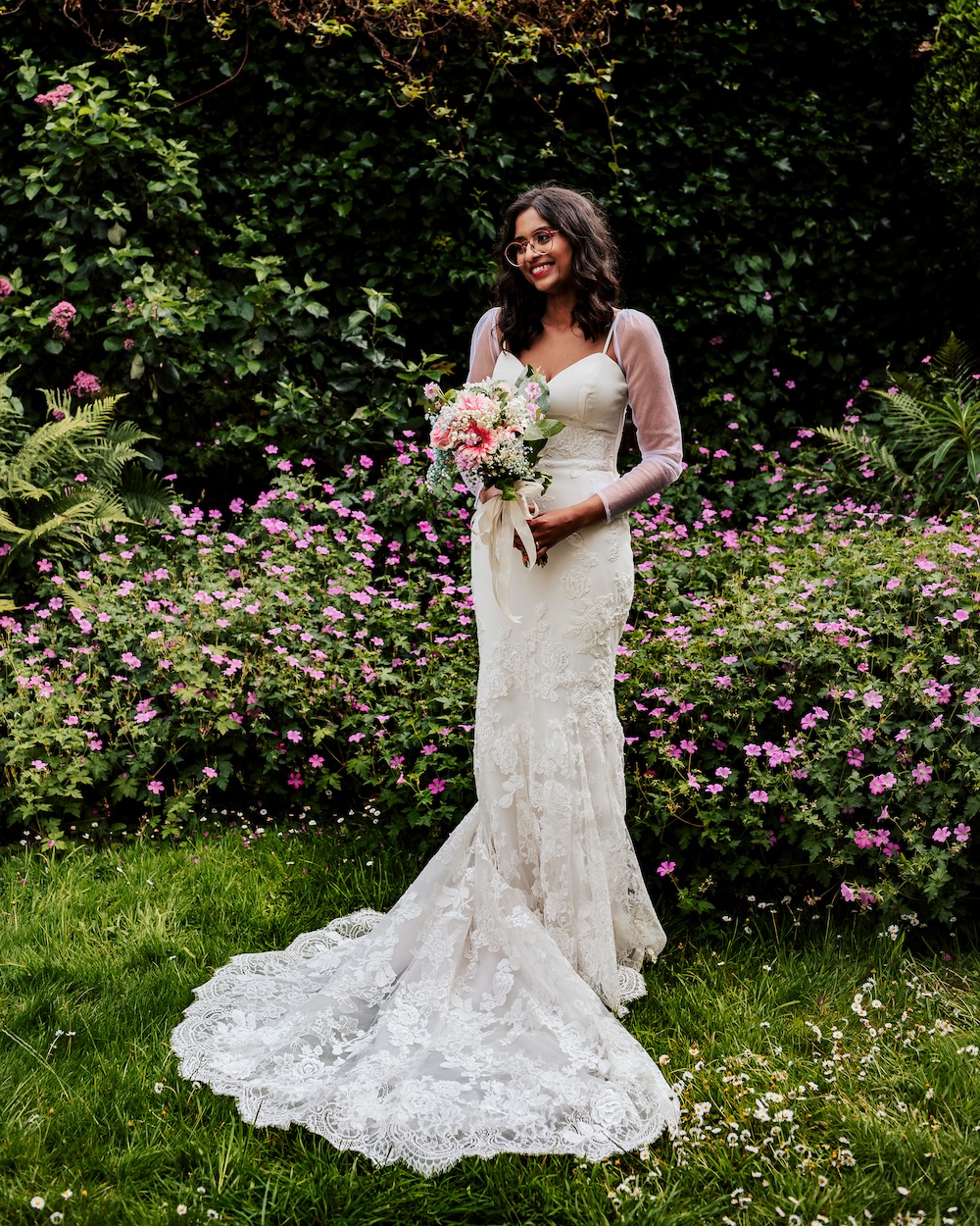 The bride stands alone in a garden at No.4 Clifton Village, a beautiful outdoor wedding venue in Bristol. She is wearing an elegant white lace gown with a long train, holding a bouquet of pastel pink and white flowers. The lush greenery and vibrant pink flowers surrounding her create a serene and romantic backdrop, showcasing the natural charm of this Clifton wedding venue, perfect for an outdoor ceremony or photoshoot.