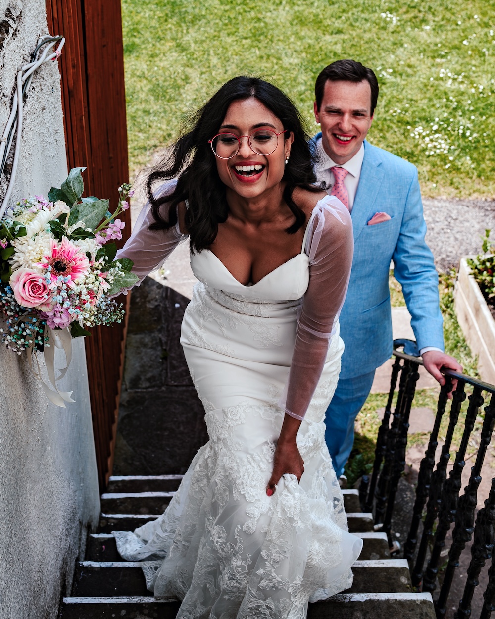 A joyful bride wearing a beautiful white wedding dress with lace details and sheer sleeves, smiles brightly while ascending the staircase from No.4 into the Secret Garden. She holds a bouquet filled with pink roses, white chrysanthemums, and baby's breath. The groom follows closely behind, dressed in a light blue suit with a pink tie, also smiling warmly. The setting is outdoors at No.4 Clifton Village, a unique wedding venue in Bristol, known for its intimate atmosphere and charming spaces. This image captures a candid moment during the couple’s wedding, showcasing the delight and excitement of the day.