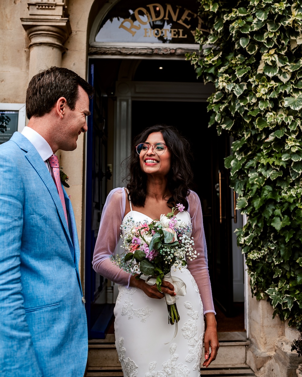 The bride and groom stand at the entrance of the Rodney Hotel in Clifton, a historic and charming part of Bristol. The Rodney Hotel is adjoined to No.4 Clifton Village, a beautiful wedding venue. The bride, holding a bouquet of pink and white flowers, smiles warmly at the groom, who is dressed in a light blue suit. The ivy-covered exterior of the hotel adds a touch of classic elegance to the scene, perfectly complementing the couple’s joyful expressions. This image captures the timeless beauty of a wedding at No.4 Clifton Village, a wedding venue with hotel.