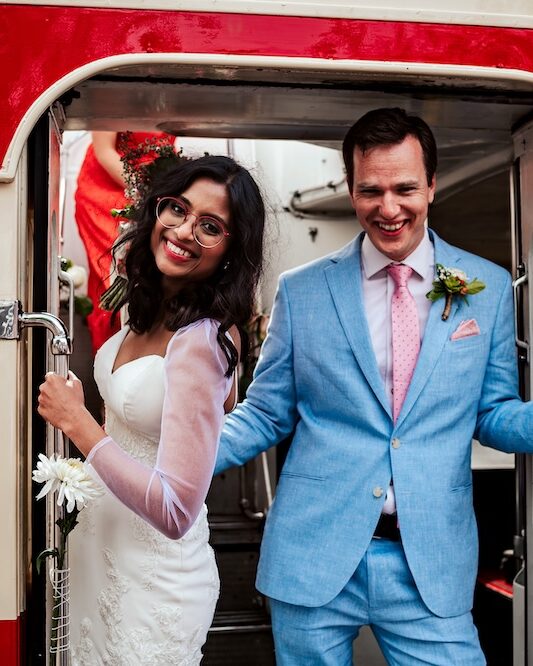 The bride and groom joyfully pose on the steps of a vintage red double-decker bus, a unique feature for wedding transportation in Bristol. The bride, in a white lace gown, and the groom, in a light blue suit, smile brightly as they stand in the doorway of the bus. This fun and distinctive element adds a playful touch to their wedding day, showcasing the couple’s personality and the versatility of hosting a wedding in Clifton.