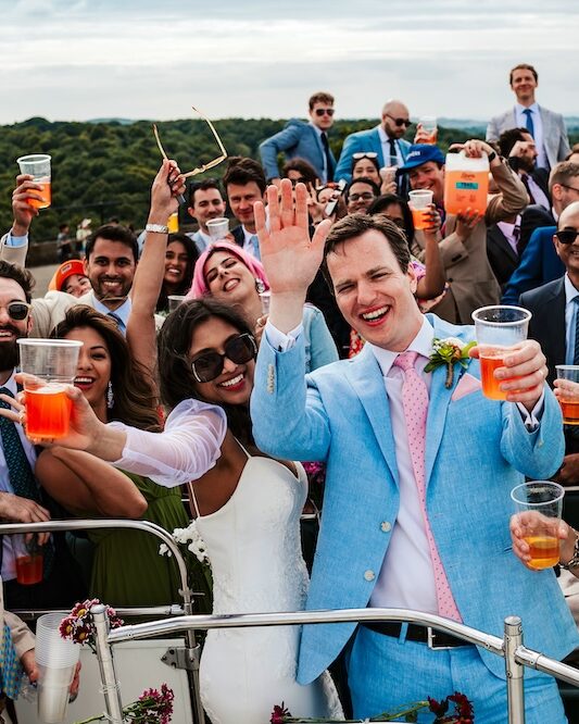 The bride, groom, and their wedding guests celebrate on the top deck of an open-top bus with drinks in hand, driving towards No.4 Clifton Village. The groom, in a light blue suit, waves enthusiastically while the bride and guests smile and cheer around him. The scenic backdrop of Bristol adds to the festive atmosphere, capturing the lively spirit of the day. This image highlights the fun and unique experiences available at a Bristol wedding, especially when hosted in the vibrant area of Clifton.