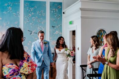 The bride and groom make their entrance into the beautifully decorated Admiral Room at No.4 Clifton Village, a popular wedding venue in Bristol. The bride, wearing a stunning white lace dress, and the groom, in a light blue suit, are greeted with smiles and applause from their guests. The room’s elegant blue wallpaper with floral patterns and the modern living chandelier centrepiece create a unique and intimate atmosphere, making it an ideal venue for a memorable wedding ceremony.