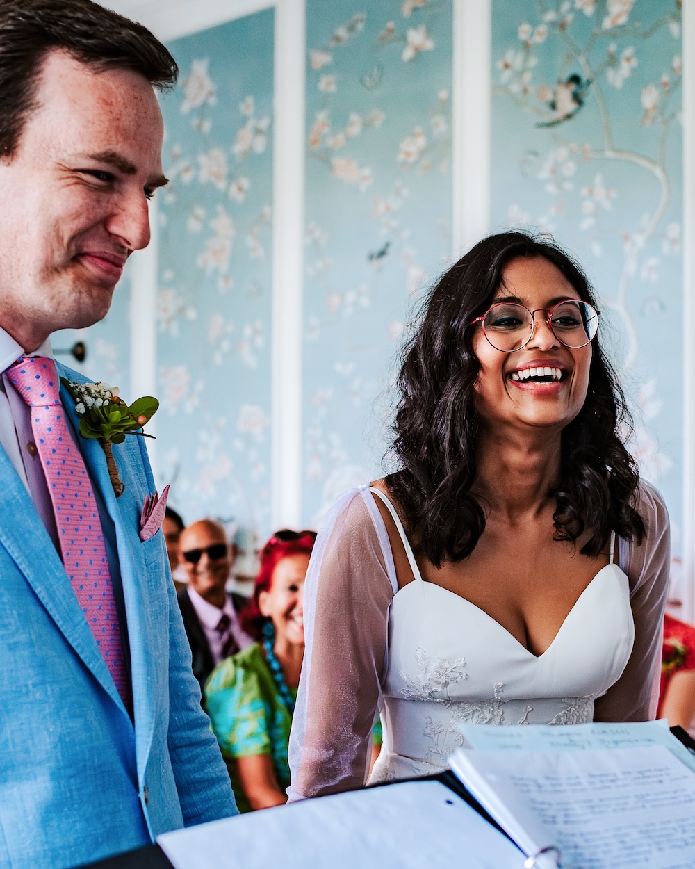 A close-up of the bride and groom during their wedding ceremony at No.4 Clifton Village, a sought-after wedding venue in Bristol. The bride beams with joy, while the groom, in a light blue suit with a pink tie, looks on with a tender smile. The backdrop of the room’s intricate blue silk wallpaper, adorned with hand-painted floral designs, adds to the charm of this intimate and unique wedding venue, perfect for couples seeking a personalized and stylish celebration.