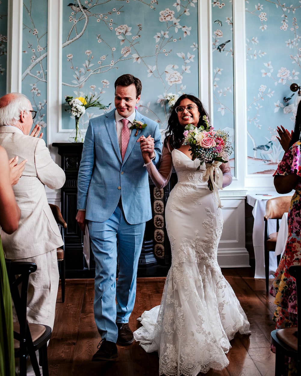 The newlyweds walk hand in hand down the aisle at No.4 Clifton Village, a charming wedding venue in Bristol, while guests applaud around them. The bride, in her fitted lace wedding gown, holds a bouquet of pink and white flowers, while the groom, in his light blue suit, looks on with pride. The room’s stunning silk wallpaper with hand-painted floral details and the original Georgian fireplace enhance the intimate and elegant ambiance of this unique wedding venue, making it a perfect choice for a small, meaningful celebration.